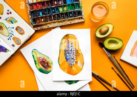 top view of avocado and papaya drawings next to colored paints, paintbrushes on yellow background Stock Photo