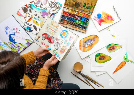 top view of woman holding album surrounded by color drawings and drawing utensils Stock Photo
