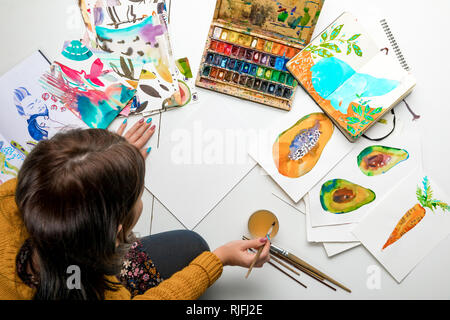 top view of woman preparing to paint with watercolors paints while surrounded by color drawings and drawing utensils Stock Photo