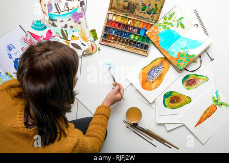 top view of woman painting with watercolors paints while surrounded by color drawings and drawing utensils Stock Photo