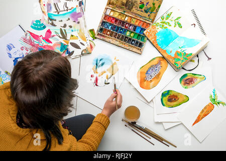 top view of woman painting with watercolors paints while surrounded by color drawings and drawing utensils Stock Photo