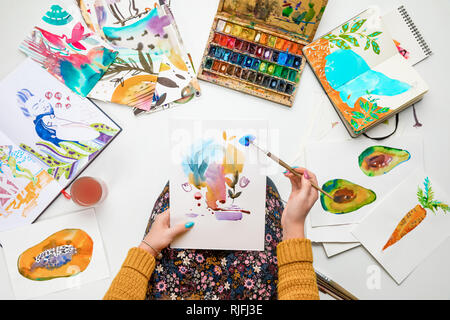 top view of woman holding drawing on knees and painting in it with watercolors paints while surrounded by colored pictures Stock Photo