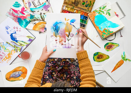 top view of woman holding drawing on knees and painting in it with watercolors paints while surrounded by colored pictures Stock Photo