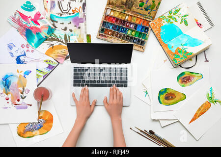 top view of female hands on laptop while surrounded by watercolor drawings Stock Photo