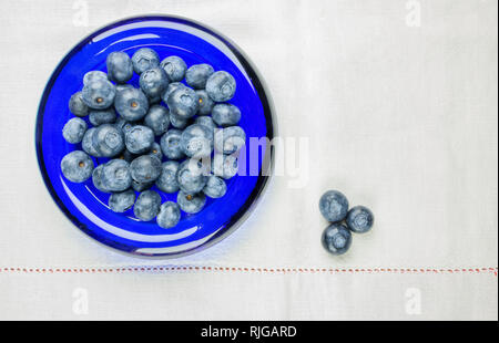 Beautiful blueberries in a blue glass bowl ,the bowl is on a white cloth , three blueberries are in the right corner of the frame Stock Photo