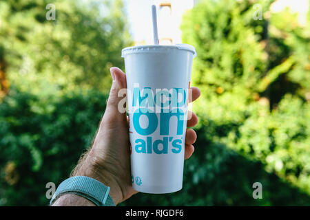 PARIS, FRANCE - JUL 27, 2018: Man holding outdoor against green background a cup of McDonald's Cola with plastic straw with blue text near one of the world's largest fast food restaurants Stock Photo