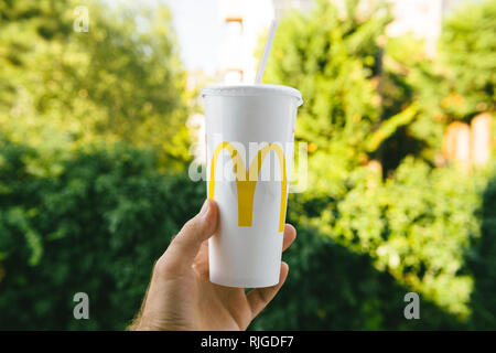 PARIS, FRANCE - JUL 27, 2018: Man holding outdoor against green background a cup of McDonald's Cola with plastic straw with yellow logo near one of the world's largest fast food restaurants Stock Photo