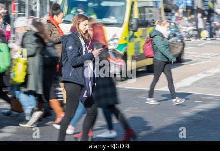 Young woman walking across a road while looking at a mobile phone, in the UK. Using smartphone while crossing road. Stock Photo