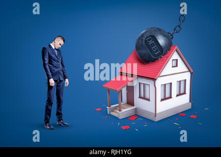 Sad businessman and metal chained ball with 'DEBT' sign breaking white private house. Financial risks. Business and commerce. Banking and finance. Stock Photo