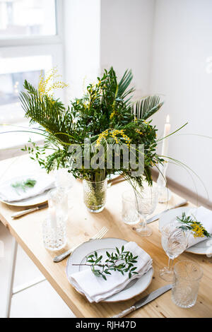 Beautiful springtime table setting with green leaves and mimosa branches, bright white table dinner decoration Stock Photo