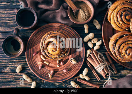 freshly baked Cinnamon rolls buns with peanuts on a earthenware plate with brown cloth, two cups of coffee and cinnamon sticks on a table. Kanelbulle  Stock Photo