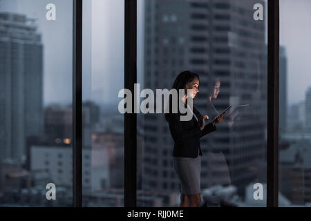 Businesswoman working late using digital tablet with city skyline in background Stock Photo