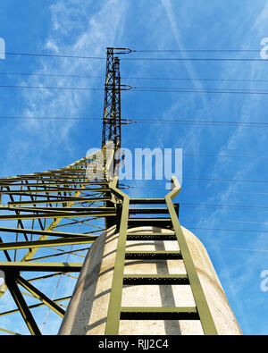 View from below up to a high voltage pylon, which stands on a concrete foundation, a ladder leads up. Above it a blue sky with light clouds of fog. Stock Photo