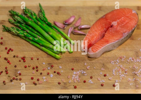 Fresh salmon steak surrounded by pink peppercorns, Himalayan salt, garlic and asparagus on a wooden cutting board, food ingredients close up Stock Photo