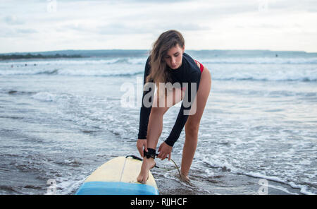 Unrecognizble barefoot woman has fixed legrope, stands on sand near surfboard, protects herself from crashing into shore lines, has active lifestyle Stock Photo