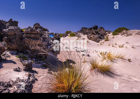 White sand dunes with rocks and bright orange grass at the coastline of Crete Island. Sea is visible through the hole in rock. Clear blue sky. Horizon Stock Photo