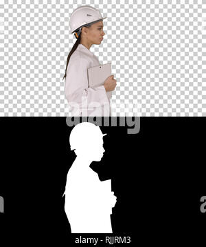 Attractive Hispanic woman in white lab coat and white safety hard hat walking holding notebook or tablet, Alpha Channel Stock Photo