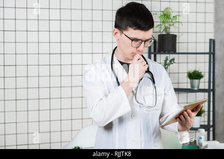Pensive doctor analyzing medical history and thinking over treatment. Handsome male practitioner holding tablet and reflecting. Digital technology in Stock Photo
