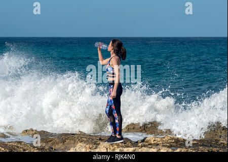sportswoman standing on rocks by the sea with crashing waves drinking from a plastic bottle of water Stock Photo