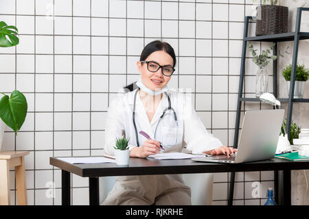 Young smiling woman sitting at desk, working on computer with medical documents in light office in hospital. Female doctor in medical gown Stock Photo