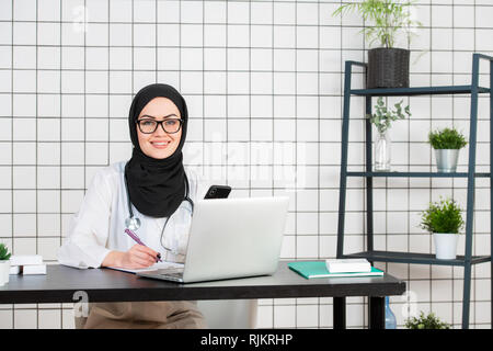 A Muslim Doctor smiling with glasses and stethoscope and documents on a white office background Stock Photo