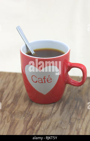 a red cup of coffee with a heart that reads: Coffee in Spanish. The table on which it is held is an old wooden board, the bottom of the image is broke Stock Photo