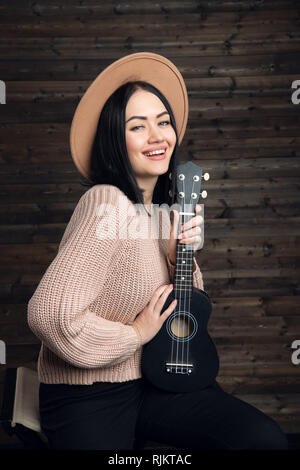 Attractive emotional young Caucasian woman in sweater and hat playing ukulele. Beautiful woman singer playing musical instrument indoors. People Stock Photo