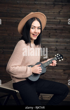 Young funny hipster girl having fun and playing on small ukulele guitar, singing and dancing. wearing vintage hat, joy, positive mood. Dark wooden Stock Photo