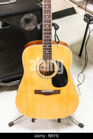 An acoustic guitar is upright on a medal stand with a black speaker behind it ready to be played. Stock Photo