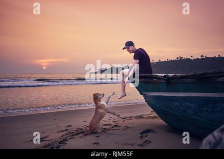 Young man relaxing on the beach and cheerful dog him giving paw at the sunrise. Tangalle in Sri Lanka. Stock Photo