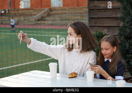 Two lovely teenage sisters have snack together next to school playground. Older girl is holding cell phone and making selfies. Stock Photo