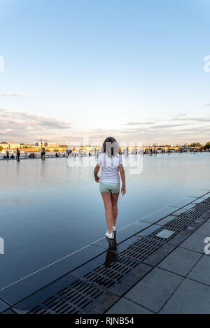Girl walking on Water Mirror  in Bordeaux. She wears white shirt. Rear view at sunset Stock Photo
