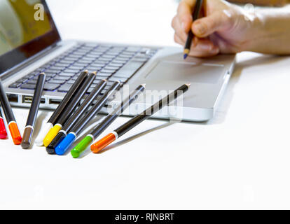 Digital or traditional graphic arts? A woman's hand seems to draw on the touch pad of a laptop computer with lots of colored pencils resting on the si Stock Photo