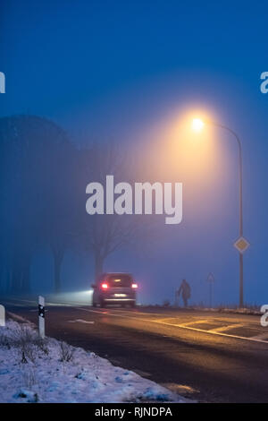 Luminous car rear lights in the night in fog on winter street with pedestrian. Stock Photo