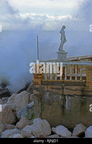 Rough sea in winter in Porto Recanati in the province of Ancona Italy with the sea crashing on rocks and a statue outside a boarded up restaurant Stock Photo