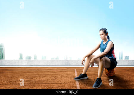 Happy asian woman with sport jersey sitting on the basketball in the outdoor basketball court Stock Photo