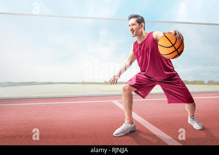 Cheerful asian basketball player man in action with the ball on the outdoor basketball court Stock Photo