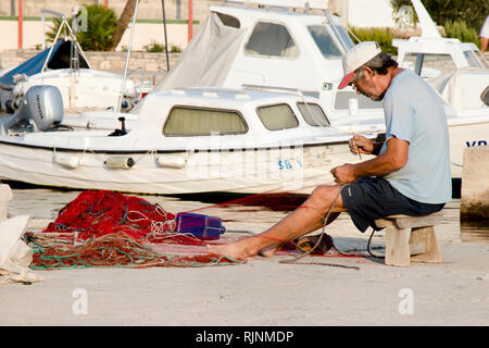 Prvic island, Croatia - August 23, 2018: Fisherman repairing his fishing net on pier with boats in the background Stock Photo