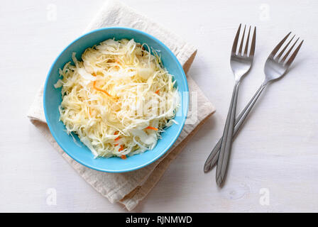 Blue bowl of shredded chopped pickled cabbage and carrot. Served with two forks on folded grey fabric towel and viewed from above on white table surfa Stock Photo