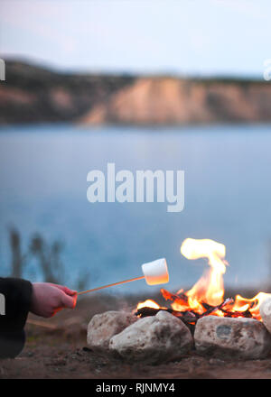 person roasting marshmallows over an open campfire in the late summer nights while camping by a lake blurred background for copy space and text overla Stock Photo