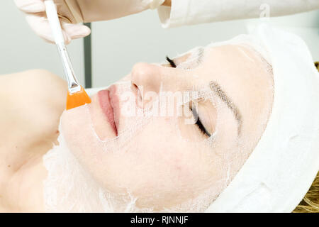 beautician worker holding paintbrush  applying facial cosmetic serum  in biological treatment to female client  in beauty salon, eyes closed, close up Stock Photo