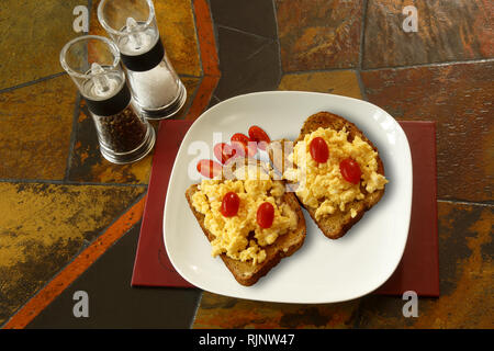 Scrambled eggs on toast with plum tomatoes. Background is a tiled table top and shows salt and pepper mills Stock Photo