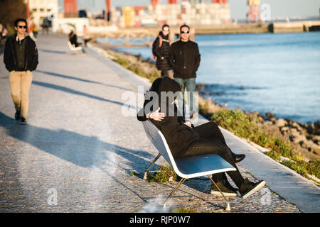 Unrecognizable people walking with side view of couple embracing on waterfront bench and observing seascape in sunlight with people walking on alley Stock Photo