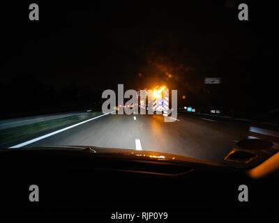 Shot from inside of car driving down road in night time with blurry lights of road signs and security traffic illuminated arrow on truck during accident or road works Stock Photo