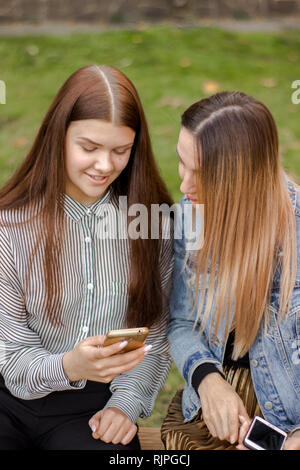 Portrait of two beautiful girls are looking into the phone, sitting on a bench in the autumn park Stock Photo