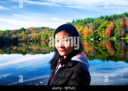 A beautiful chinese woman looking at the camera with a fall foliage reflecting in the water background in autumn at Burr Pond state park in torrington Stock Photo