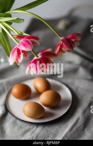 Boiled brown eggs on a plate next to the tulips in a transparent glass cup on a gray background in a muffled light and in soft focus Stock Photo