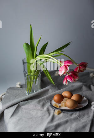 Boiled brown eggs on a plate next to the tulips in a transparent glass cup on a gray background in a muffled light and in soft focus Stock Photo