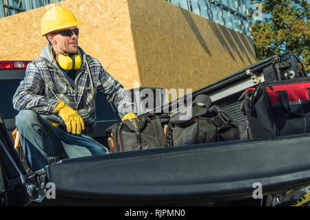 Smiling Caucasian Construction Worker Seats on a Pickup Truck. Men Wearing Yellow Hard Hat, Safety Glasses and Gloves. Stock Photo