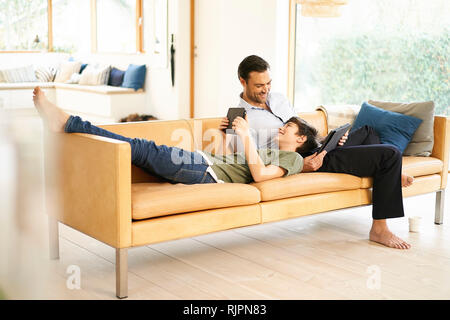 Boy and father using digital tablets whilst relaxing on sofa Stock Photo